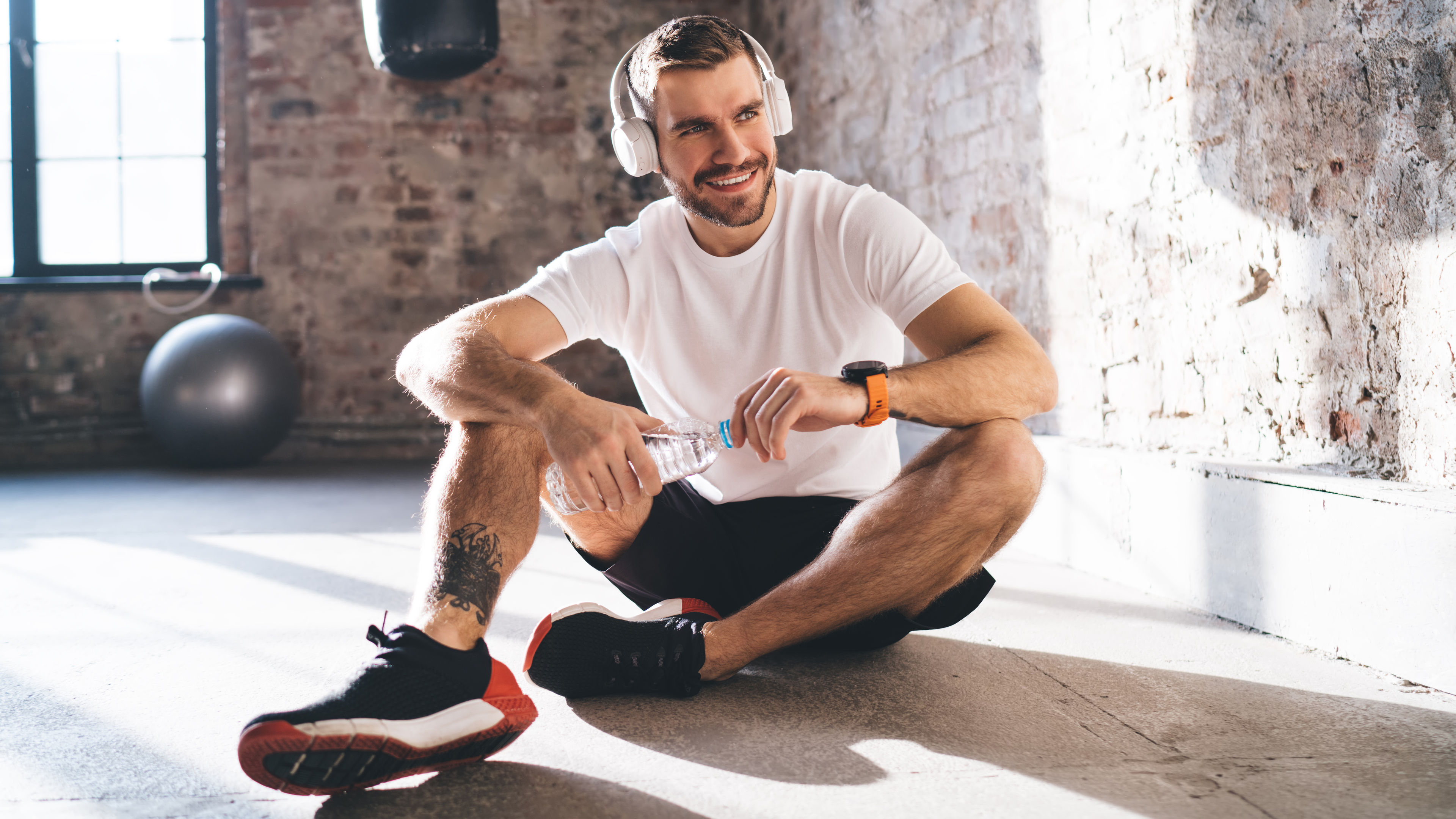 A sportsman sitting on the floor with headphones on, holding a bottle of drinking water in his hands.