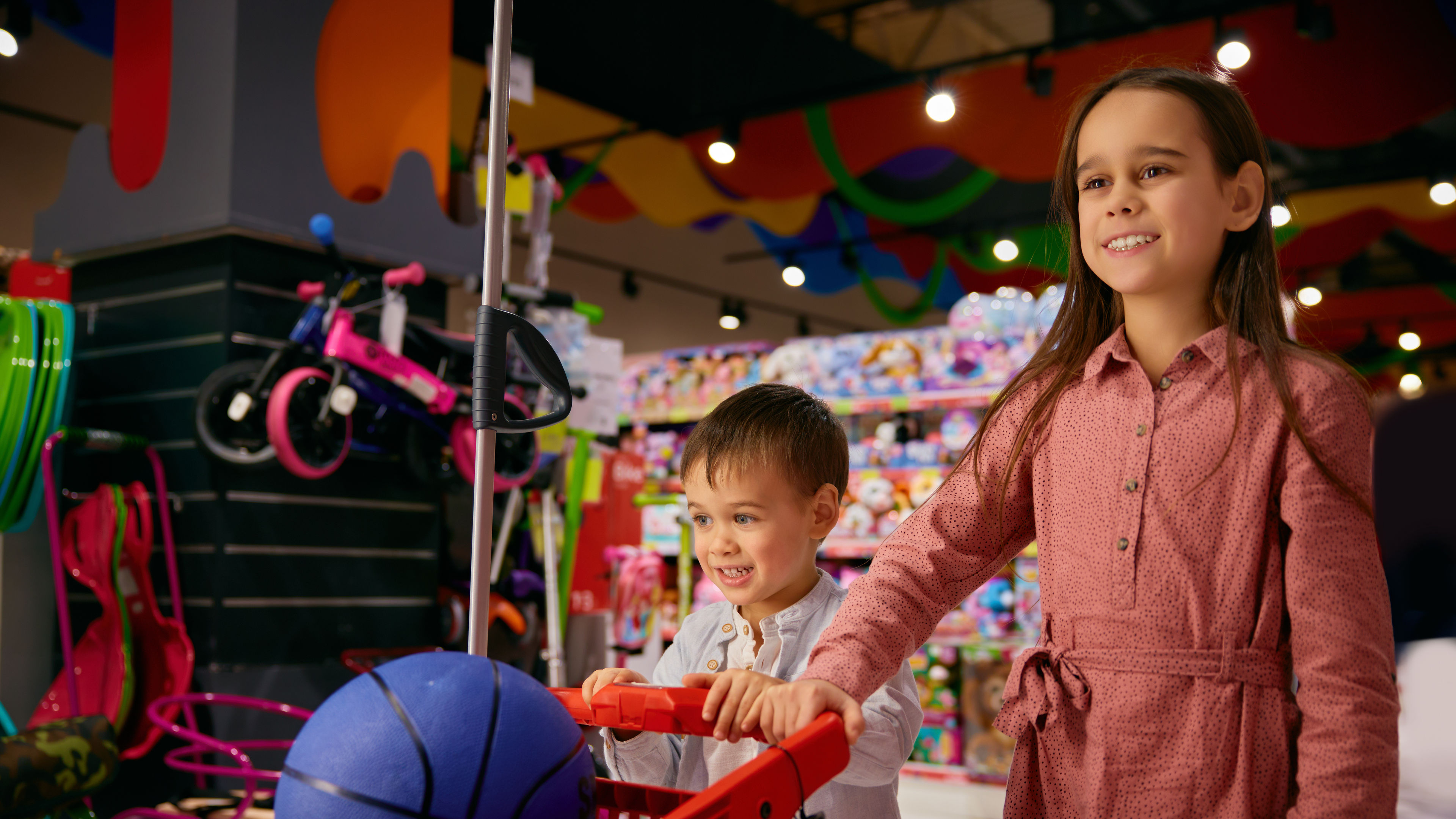 Two little kids pushing a shopping cart that has a football inside it while exploring the aisles of the store.