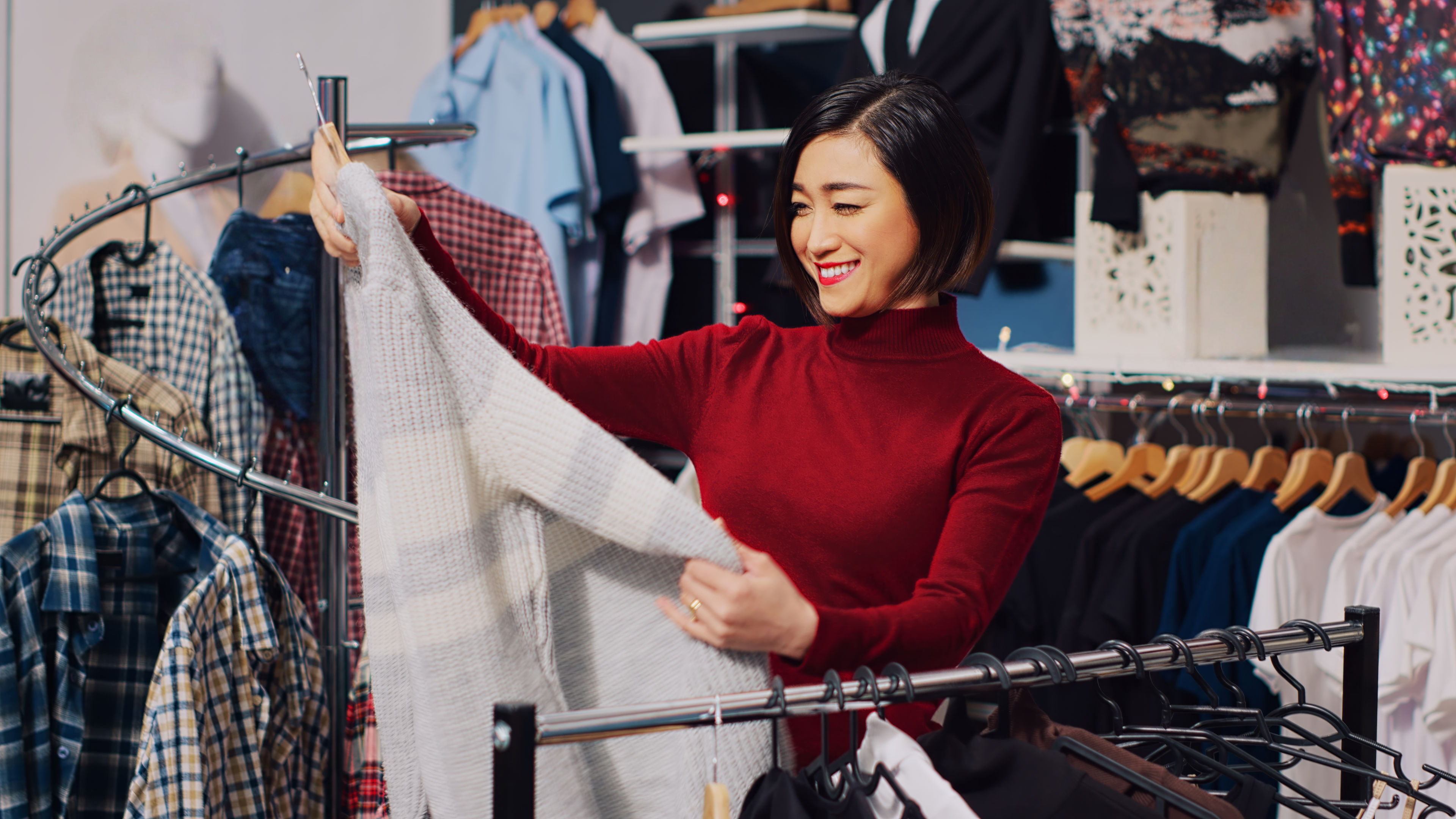A woman in a red turtleneck sweater smiles while shopping, holding up a soft plaid garment in a clothing store.