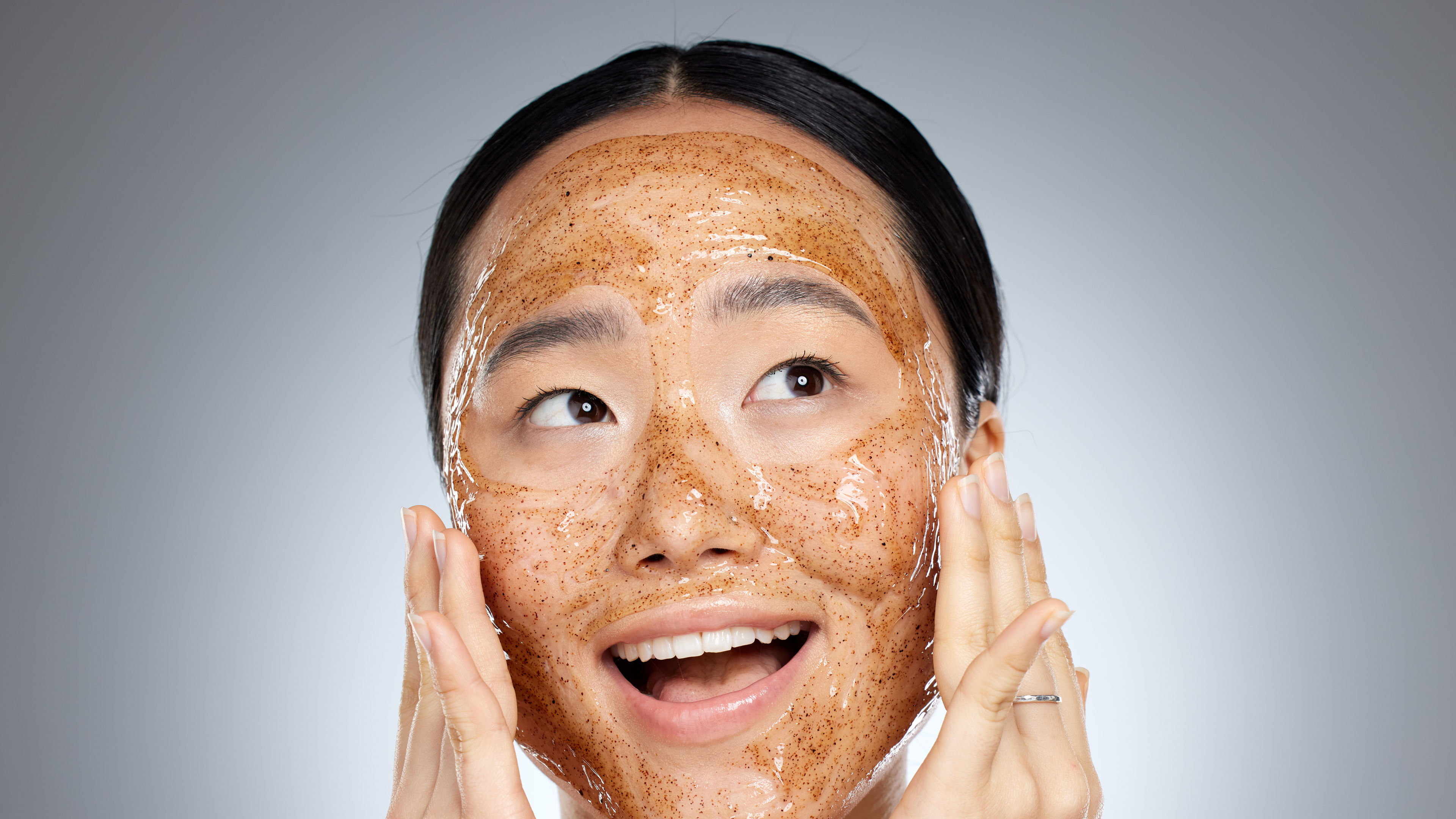 A woman is smiling while applying an exfoliating face mask on her skin. 