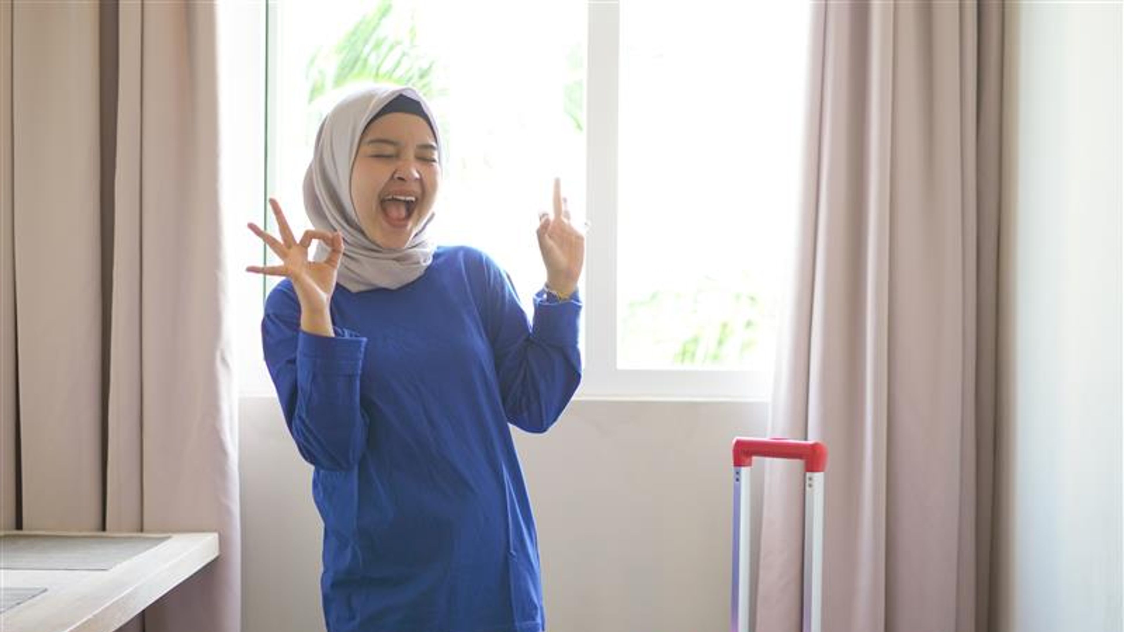 asian hijab woman cheerful wearing blue t-shirt and raising hand with okay gesture in a hotel.