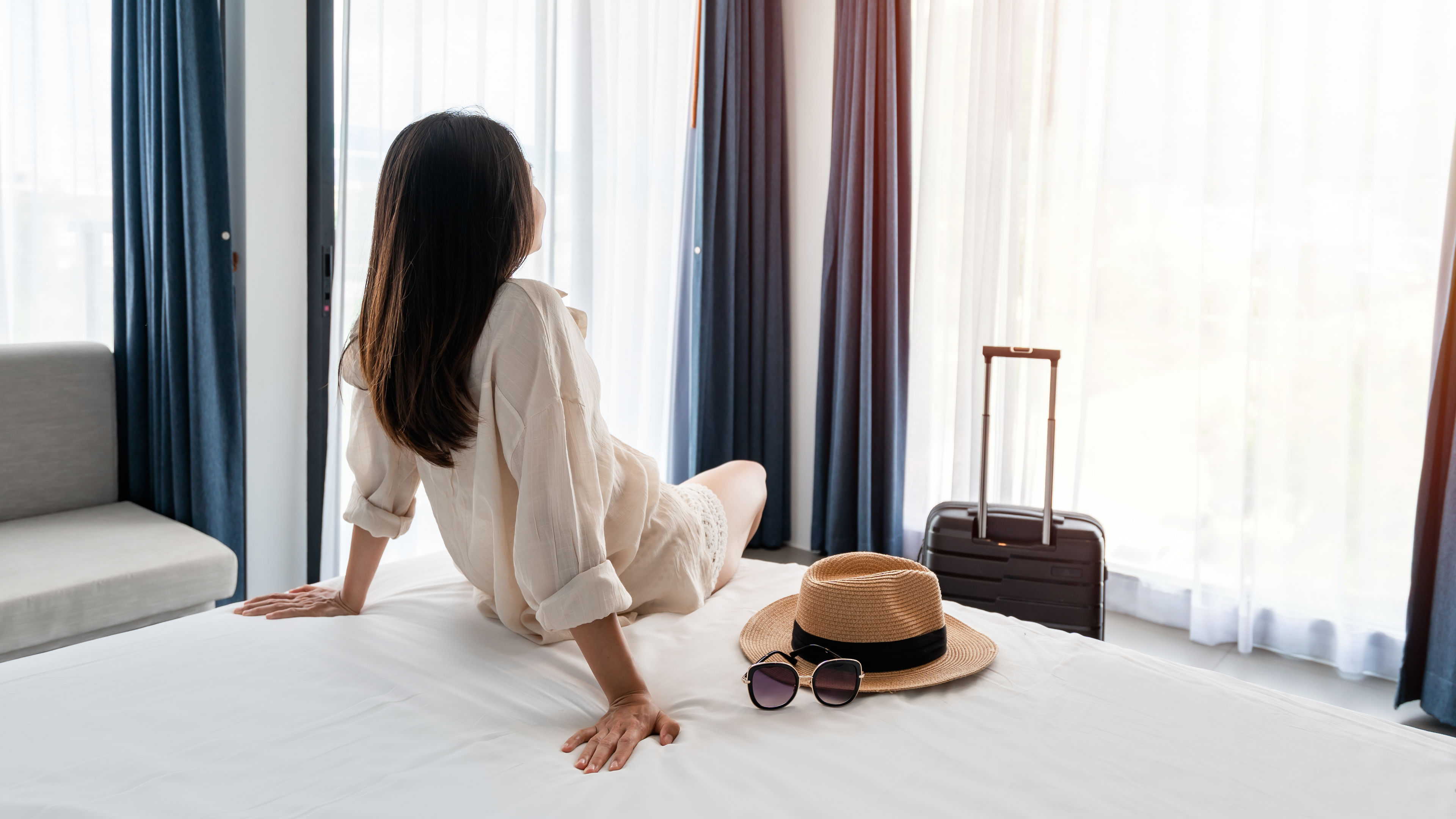 Young asian woman traveler with luggage sitting and relaxing on the bed in hotel room 