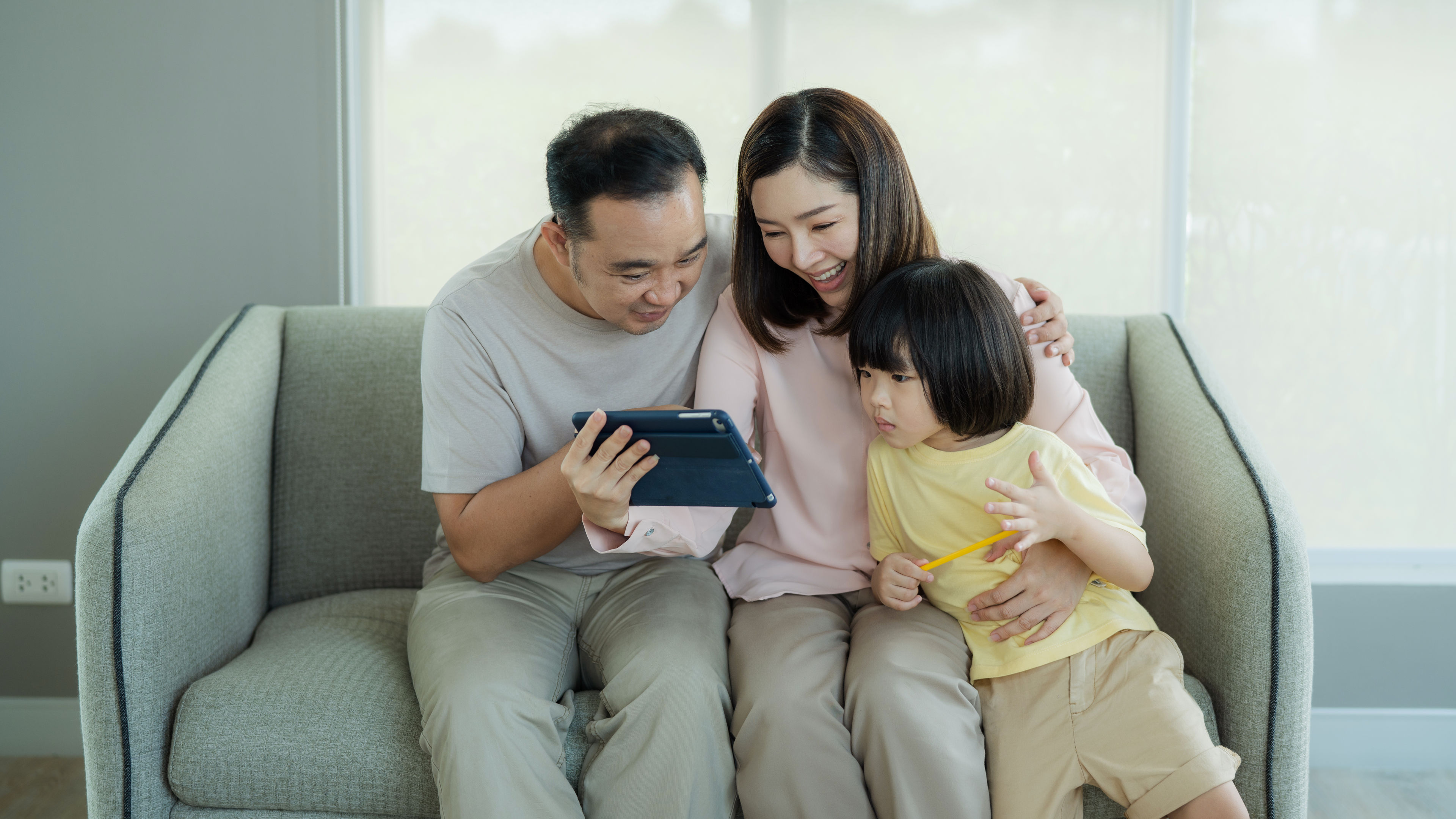 A happy family sits together on a couch, smiling and enjoying content on a tablet.  