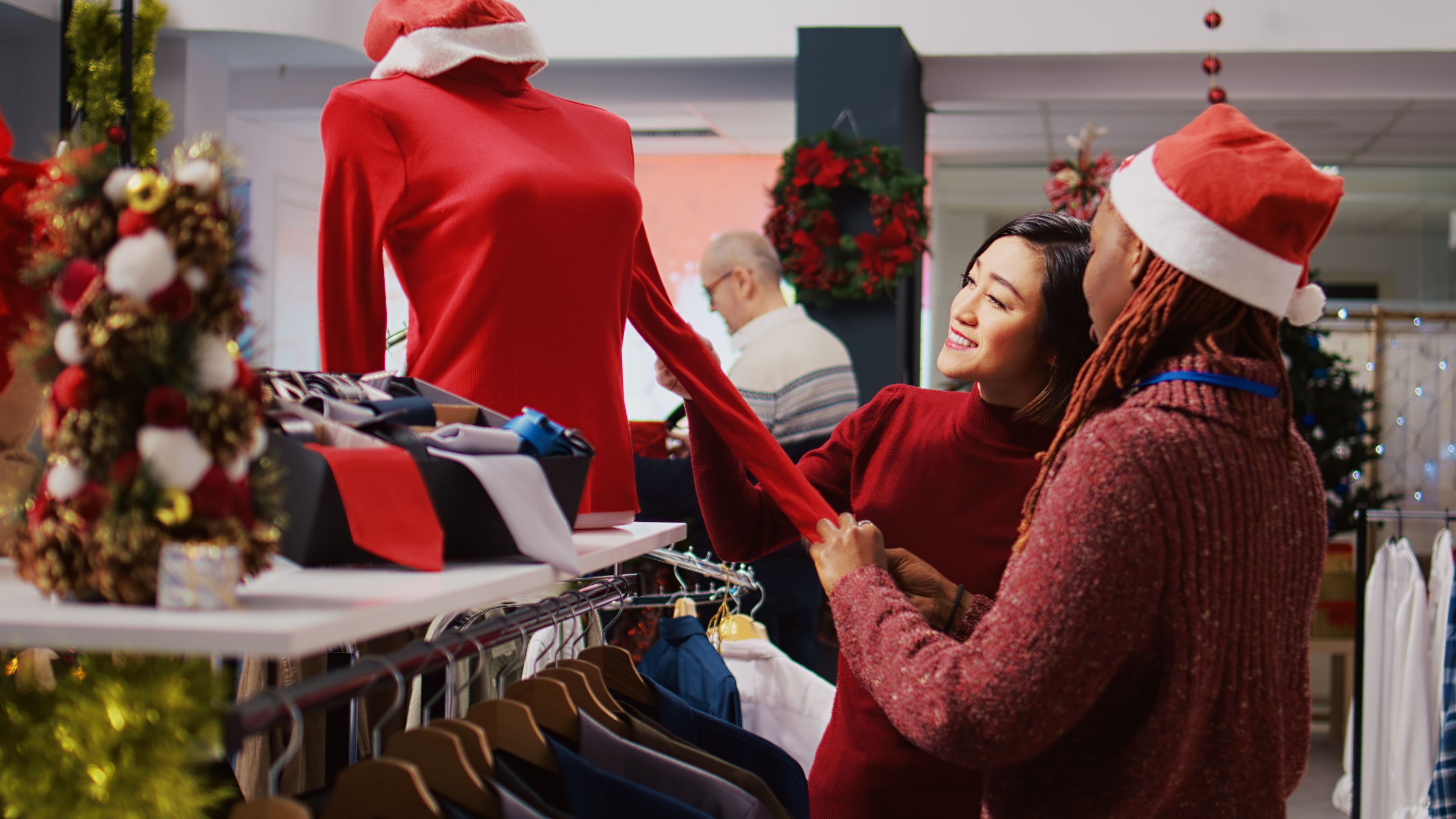 Two people in festive outfits are shopping for holiday clothing and looking at a red sweater. 
