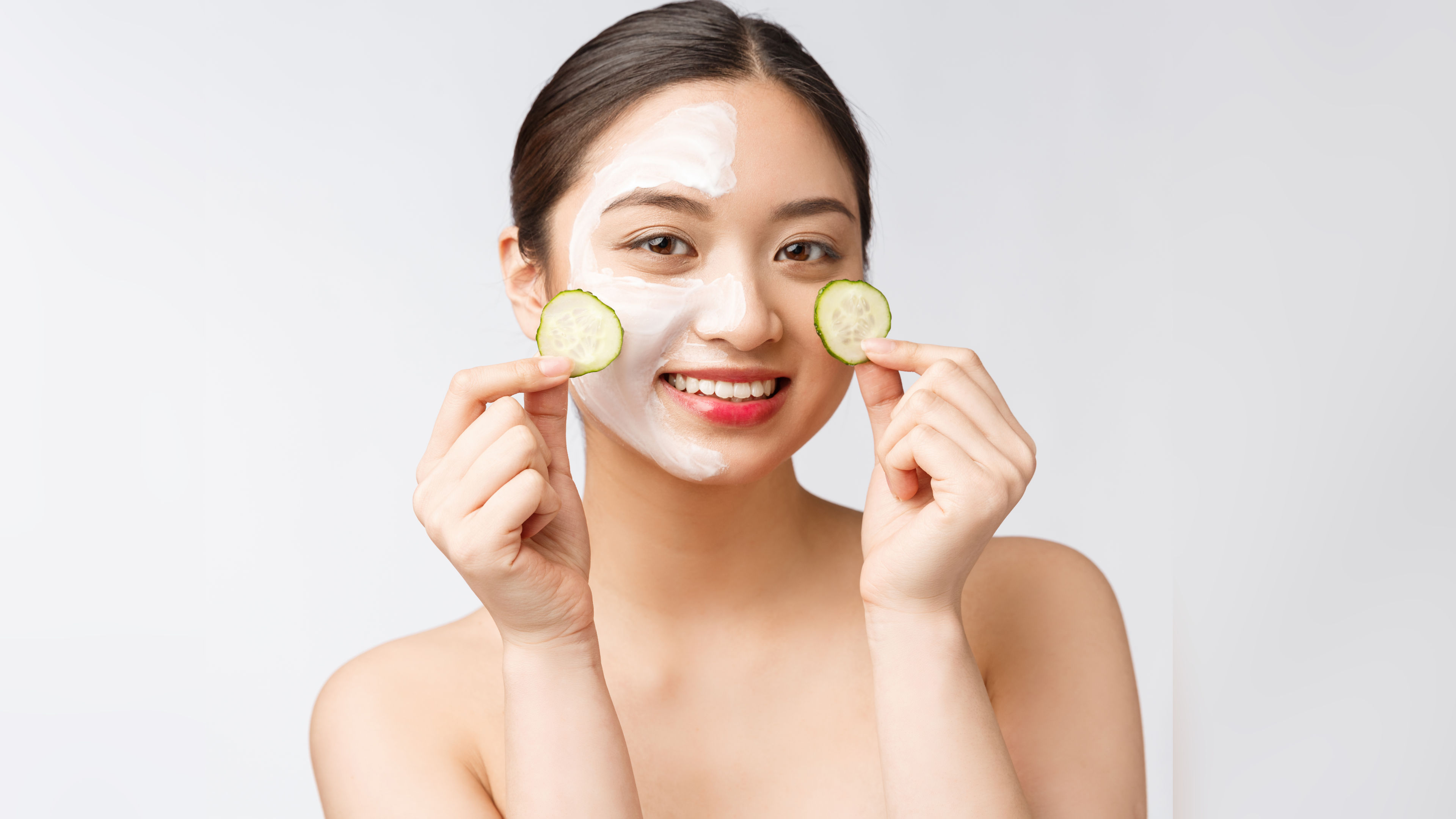 A woman applies a face mask while holding cucumber slices for skincare. 